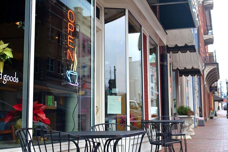 A neon OPEN sign with a steaming mug of coffee in the window of a cafe with outdoor seating.